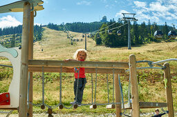 Kleines Kind auf Spielgerüst mit Blick auf Bergbahn und Wiesenhang.