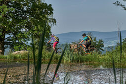 Zwei Mountainbiker fahren am Teich vorbei mit Blick auf hügelige Landschaft.