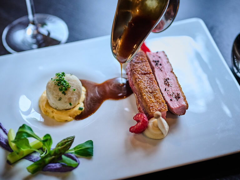 Entenbrust mit Sauce, Kartoffelknödel und Gemüse auf weißem Teller angerichtet.