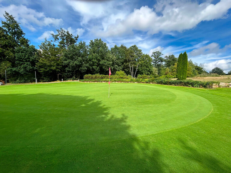 Sonnige Golfanlage mit Fahne, umgeben von Bäumen und gepflegtem Grün.