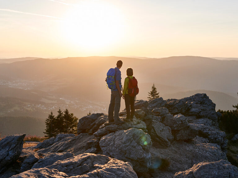 Wanderpaar mit Rucksäcken auf Felsen blickt bei Sonnenuntergang ins Tal.
