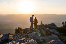 Wanderpaar mit Rucksäcken auf Felsen blickt bei Sonnenuntergang ins Tal.